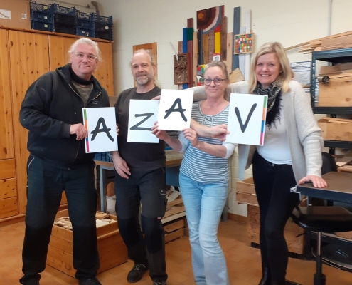 Vier Personen im Berufsbildungsbereichs der Stralsunder Werkstätten halten Schilder mit der Buchstabenkombination AZAV in den Händen.