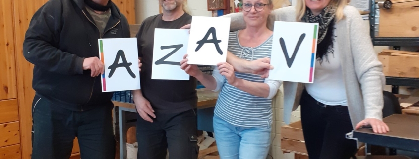 Vier Personen im Berufsbildungsbereichs der Stralsunder Werkstätten halten Schilder mit der Buchstabenkombination AZAV in den Händen.