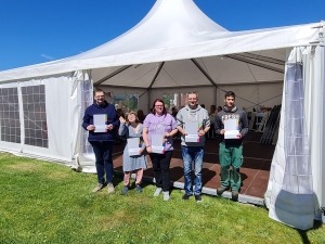 Fünf Personen stehen vor einem Pavillon. Sie halten ein Zertifikat in ihren Händen.