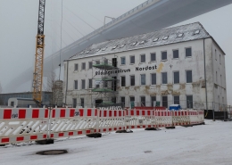 Die neue Betriebsstätte der Stralsunder Werkstätten mitten im Baugeschehen befindet sich im dichten Nebel. Im Vordergrund sind Bauzäune, ein Kran sowie ein Baugerüst zu sehen.