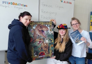 Drei Personen zeigen ein fertiges Puzzle mit Dinosauriermotiv und ein Fachbuch.