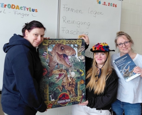 Drei Personen zeigen ein fertiges Puzzle mit Dinosauriermotiv und ein Fachbuch.