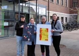 Vier Personen stehen vor dem Helios Hanseklinikum in Stralsund und halten zwei Bilder der Wanderfotoausstellung der Stralsunder Werkstätten in den Händen.