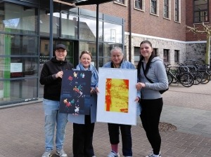 Vier Personen stehen vor dem Helios Hanseklinikum in Stralsund und halten zwei Bilder der Wanderfotoausstellung der Stralsunder Werkstätten in den Händen.