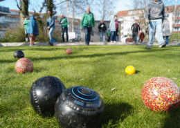 Im Vordergrund liegen verschiedenfarbige Kugeln während eines Rasenbowlingspiels. Im Hintergrund sind viele Spieler zu sehen.