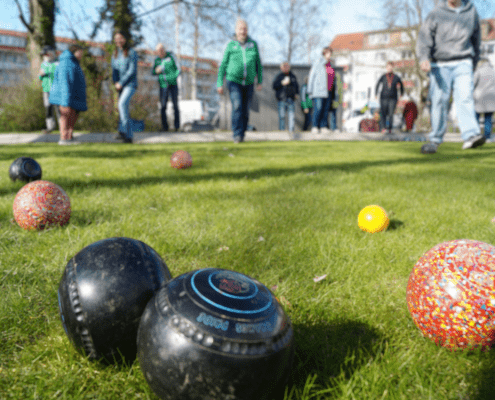 Im Vordergrund liegen verschiedenfarbige Kugeln während eines Rasenbowlingspiels. Im Hintergrund sind viele Spieler zu sehen.