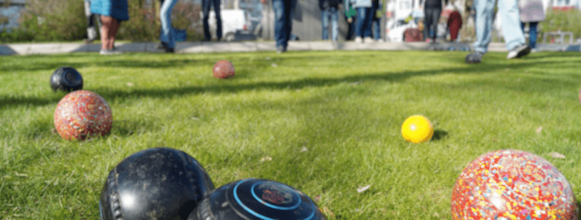 Im Vordergrund liegen verschiedenfarbige Kugeln während eines Rasenbowlingspiels. Im Hintergrund sind viele Spieler zu sehen.