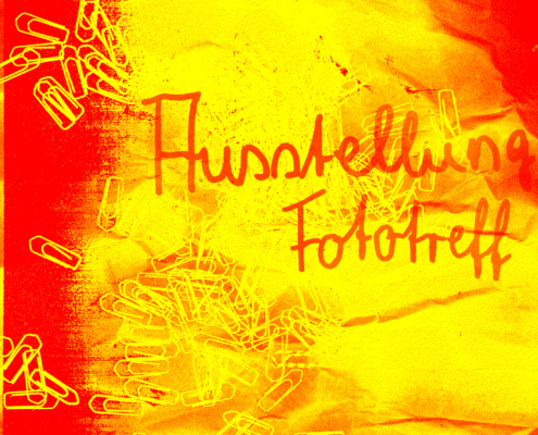 Ein Plakat in rot, orange und gelb mit der Aufschrift "Ausstellung Fototreff". Im Hintergrund sind Büroklammern zu sehen.