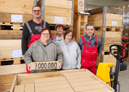 Fünf Personen stehen hinter einer voll beladenen Holzkiste. Eine Person hält ein Paket mit der Aufschrift "7.000.000" in den Händen.