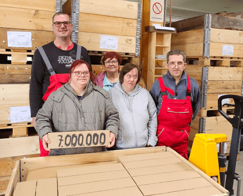 Fünf Personen stehen hinter einer voll beladenen Holzkiste. Eine Person hält ein Paket mit der Aufschrift "7.000.000" in den Händen.
