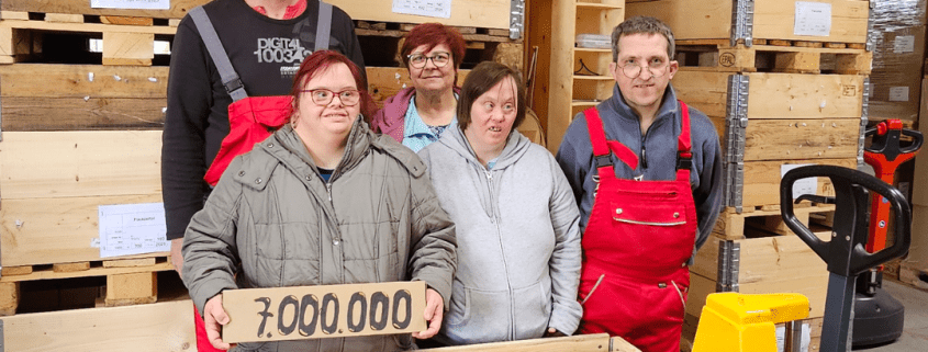 Fünf Personen stehen hinter einer voll beladenen Holzkiste. Eine Person hält ein Paket mit der Aufschrift "7.000.000" in den Händen.
