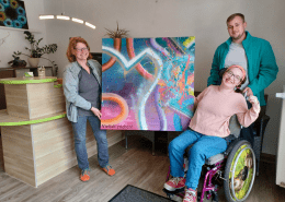 Eine Person übergibt eine große farbenfroh gestaltete Leinwand an einer weitere Person. Das Kunstwerk trägt die Aufschrift Vielfalt erleben.