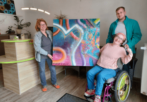 Eine Person übergibt eine große farbenfroh gestaltete Leinwand an einer weitere Person. Das Kunstwerk trägt die Aufschrift Vielfalt erleben.