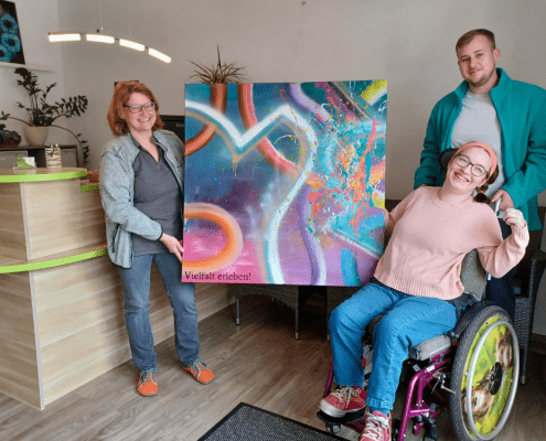 Eine Person übergibt eine große farbenfroh gestaltete Leinwand an einer weitere Person. Das Kunstwerk trägt die Aufschrift Vielfalt erleben.