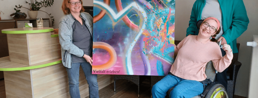 Eine Person übergibt eine große farbenfroh gestaltete Leinwand an einer weitere Person. Das Kunstwerk trägt die Aufschrift Vielfalt erleben.