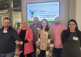 Sechs Personen der Stralsunder Werkstätten stehen vor dem Willkommensschild zum Inklusiven Netzwerktreffen am 18. März 2025.