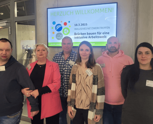 Sechs Personen der Stralsunder Werkstätten stehen vor dem Willkommensschild zum Inklusiven Netzwerktreffen am 18. März 2025.