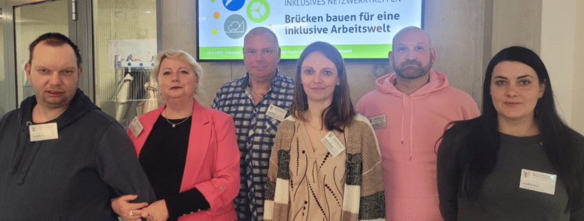 Sechs Personen der Stralsunder Werkstätten stehen vor dem Willkommensschild zum Inklusiven Netzwerktreffen am 18. März 2025.