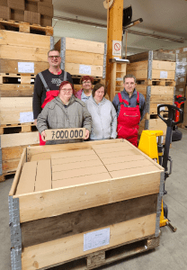 Fünf Personen stehen hinter einer voll beladenen Holzkiste. Eine Person hält ein Paket mit der Aufschrift "7.000.000" in den Händen.