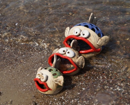 drei Keramikfische mit großem offenen Mund in unterschiedlichen Größen am Strand