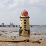 ein Keramikleuchtturm am Strand, im Hintergrund sieht man die Stadt Stralsund