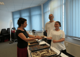 Zwei Personen des Berufsbildungsbereichs servieren einem Gast ein Stück Kuchen.