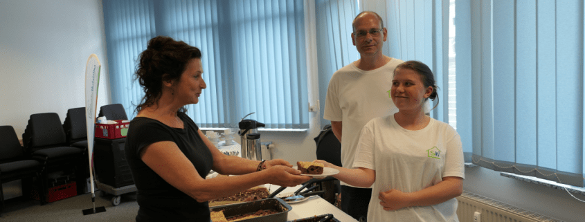 Zwei Personen des Berufsbildungsbereichs servieren einem Gast ein Stück Kuchen.