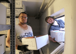 Zwei Personen haben Bücherkisten in den Händen. Eine Person schaut dabei durch ein Fenster.