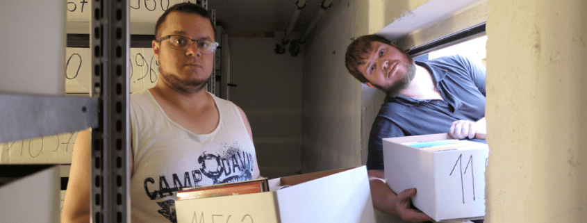 Zwei Personen haben Bücherkisten in den Händen. Eine Person schaut dabei durch ein Fenster.