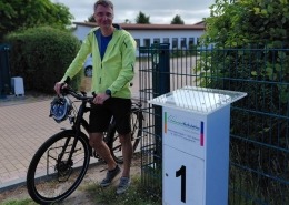 Ein Mann steht mit seinem Fahrrad neben einem Schild, auf dem die Zahl 1 abgebildet ist.